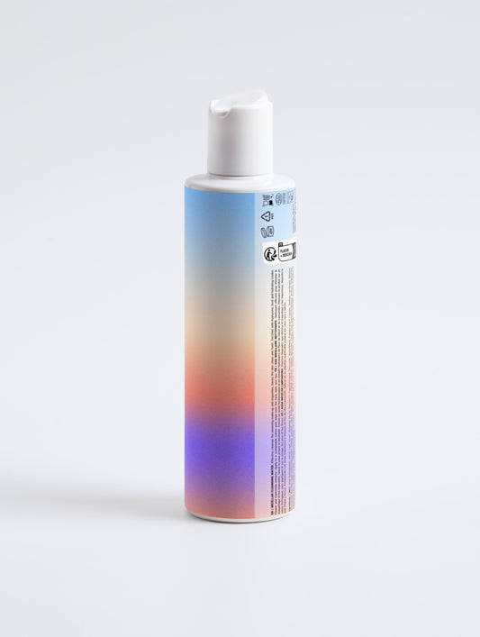 A bottle with a pump dispenser and a gradient label in blue, pink, orange, and purple stands upright on a white background, ideal for highlighting Micellar Cleansing Water, a gentle makeup remover and cleanser.